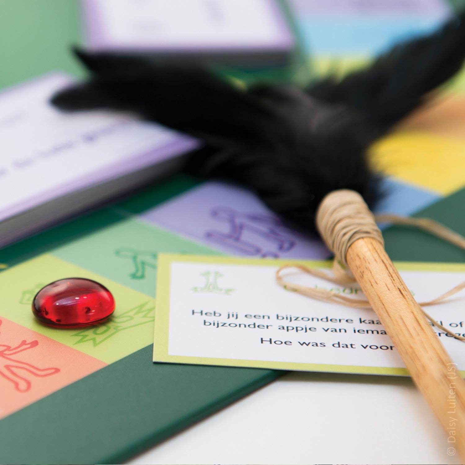 Een hoek van het groene spelbord Ons Bestaan Op Een Bordje met een kleurig traject is zichtbaar met in de achtergrond twee paarse stapels kaarten. Op de voorgrond ligt in het kleurige traject een rode glazen traan op een groen vlakje. Alsof wij (de kijkers) het net zelf hebben neergelegd om even na te denken, ligt over het traject een houten praatstok met zwarte veren, met daarbij een groene vragenkaart. Op deze vragenkaart uit de categorie 'Gebeurtenis' staat de vraag: Heb jij een bijzondere kaart, email of een bijzonder appje van iemand gekregen? Hoe was dat voor jou?