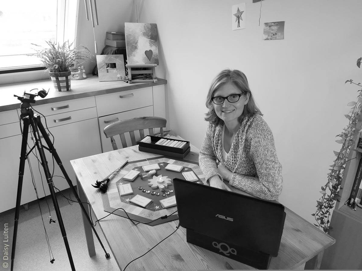 In een zwartwit afbeelding zien we Daisy Luiten aan tafel zitten met haar therapeutische tool uitgestald. Een camera op een statief staat op het spel gericht. Daisy heeft ook haar laptop voor zich waarop ze via beeldbellen online sessies geeft over het werken met Ons Bestaan Op Een Bordje. De therapeutische tool die vertellen over een nare of pijnlijke ervaring vergemakkelijkt.