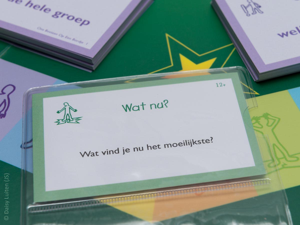 Onderdeel_Ons-Bestaan-Op-Een-Bordje_Vragenkaart_Categorie_Wat-nu_1_Daisy-Luiten_1200x900 Een hoek van het groene spelbord Ons Bestaan Op Een Bordje met een kleurig traject is zichtbaar met in de achtergrond twee paarse stapels kaarten. Op het kleurige traject ligt een vragenkaart in een doorzichtig plastic mapje. Op deze vragenkaart uit de categorie 'Wat nu?' staat de vraag: Wat vind je nu het moeilijkste?